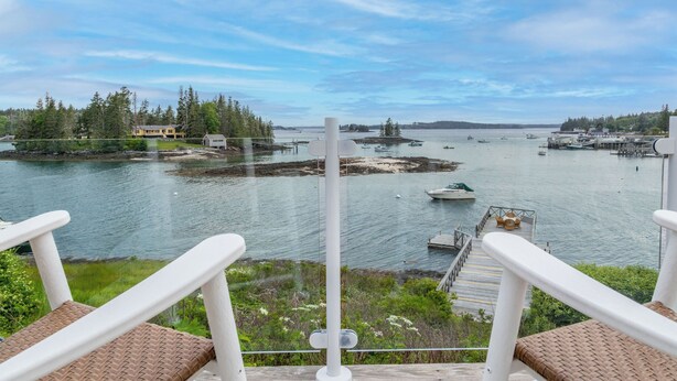 A view from a balcony overlooking a marina with boats, a dock, and a forested island. - Timeless Days, Oceanfront Fisherman’s Cottage, Deep Water Dock & Boathouse (Port Clyde)