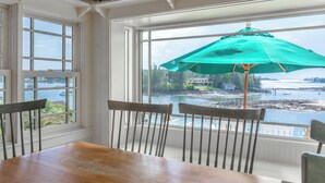 A dining area with a table and chairs, a large window with a view of a body of water, and a green umbrella. - Timeless Days, Oceanfront Fisherman’s Cottage, Deep Water Dock & Boathouse (Port Clyde)