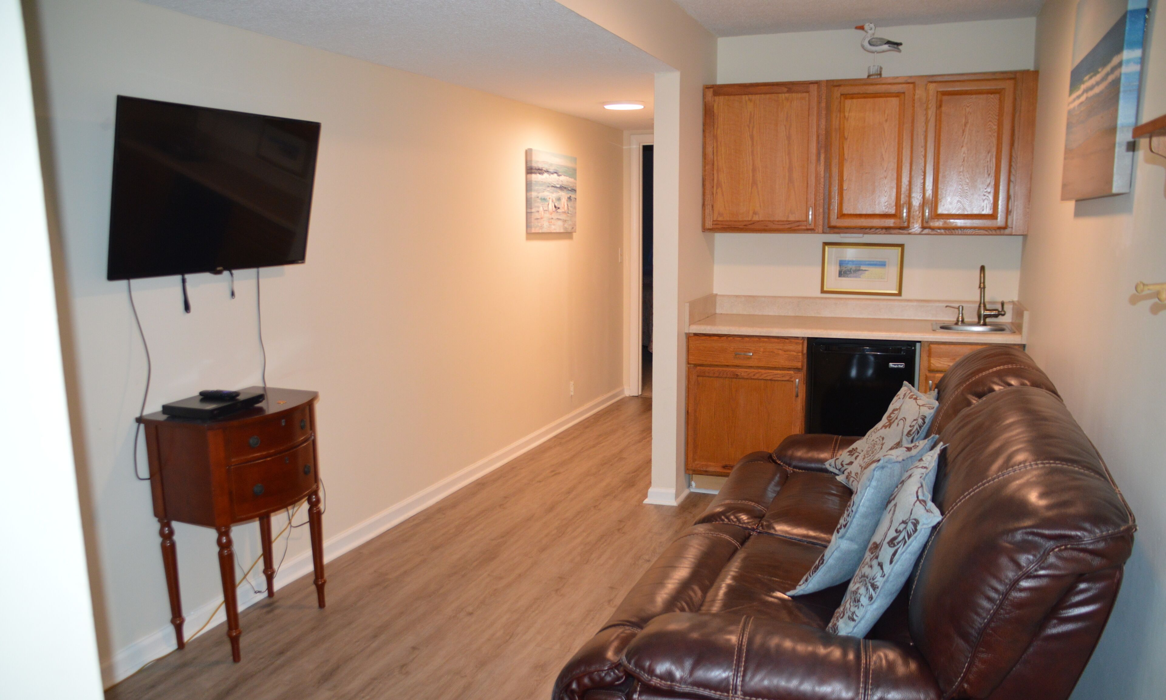 A living room with a leather couch, a television mounted on the wall, wooden cabinets, and a kitchen area with a dishwasher.