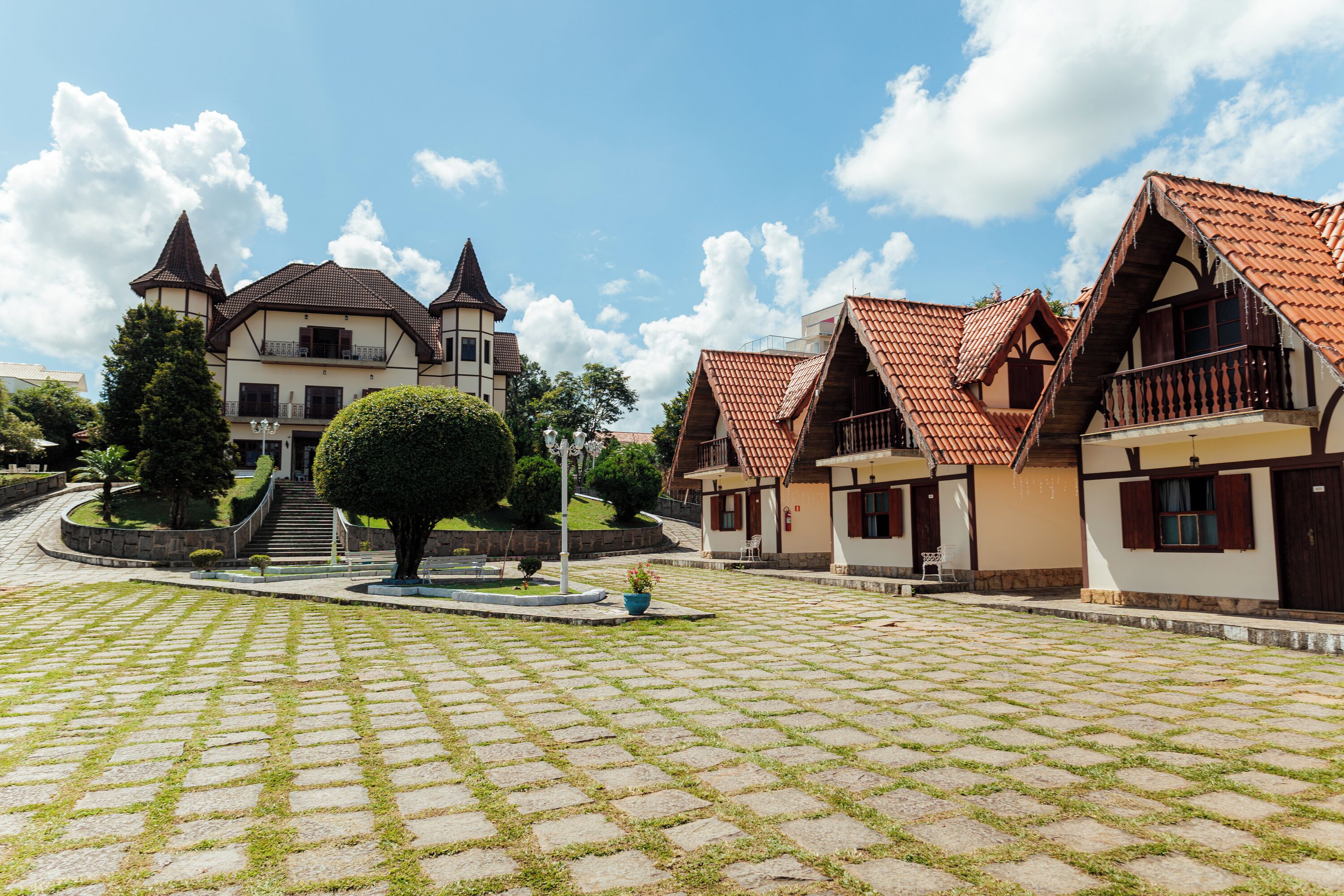 A courtyard with cobblestone paving, traditional European-style buildings with pitched roofs, and a central tree.