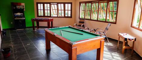 A pool table in a room with large windows, a foosball table, and an arcade machine.