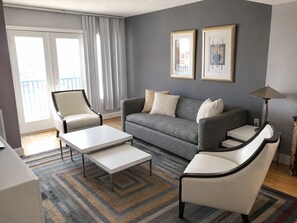 A modern living room with a grey sofa, two armchairs, a coffee table, and a floor lamp. There are two framed artworks on the wall, a sliding glass door, and a patterned rug.