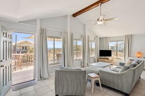 Ein großzügiges Wohnzimmer mit einer großen Couch, einem Couchtisch und einem Flachbildfernseher. Ein Balkon bietet Blick auf das Meer und die Gebäude.