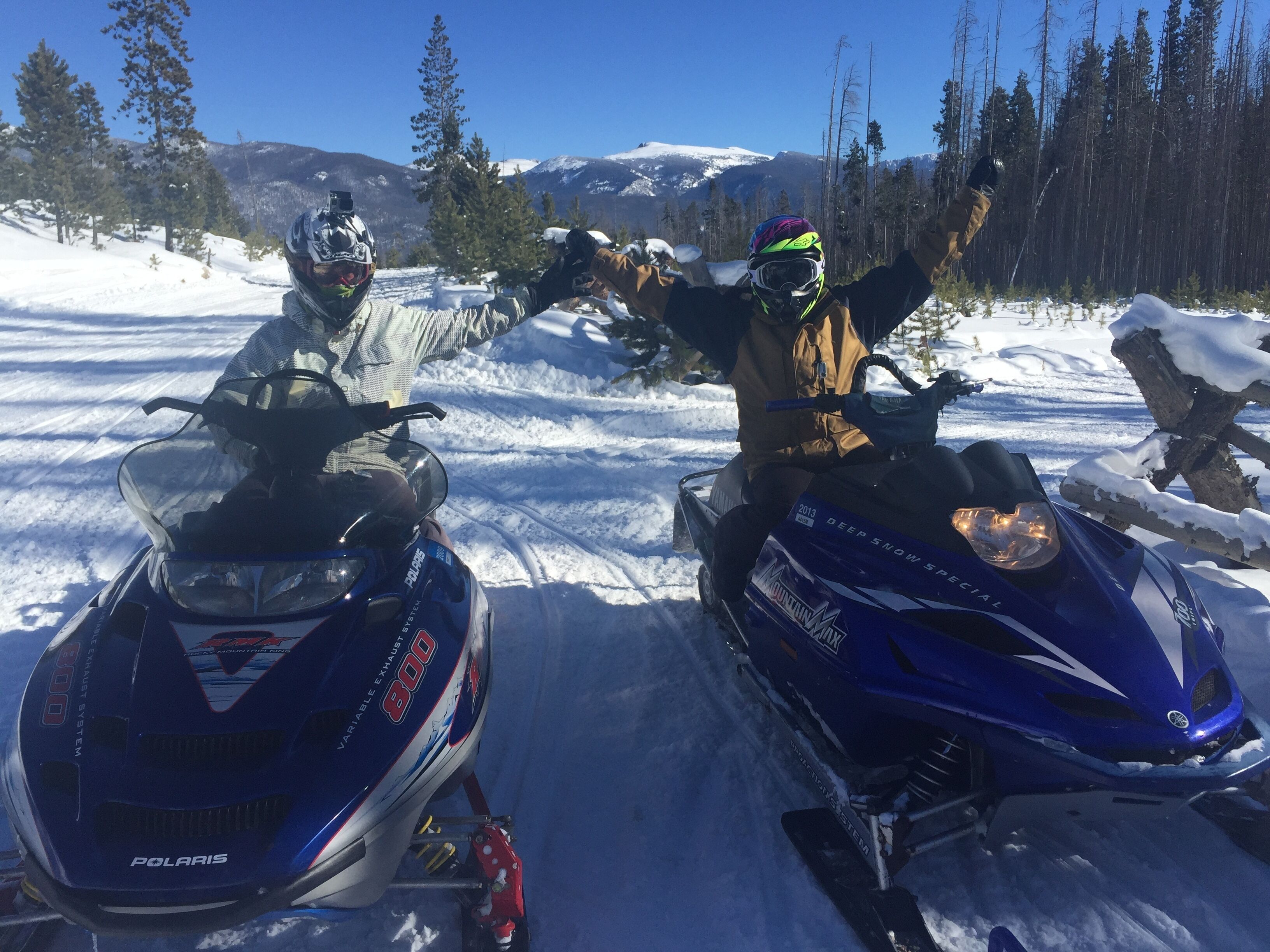 Two snowmobiles on a snowy path with a forest and mountains in the background.
