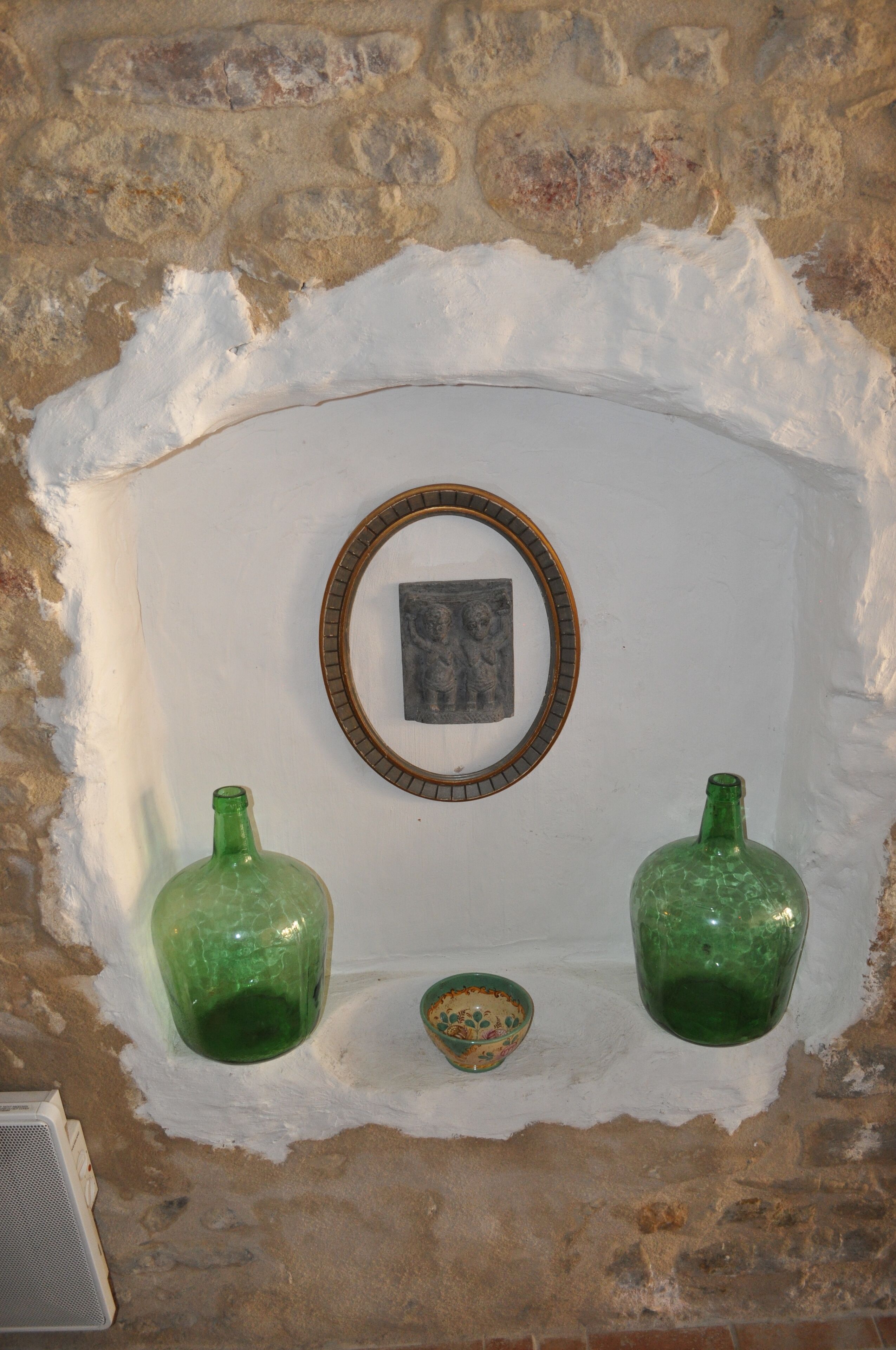 Un mur avec une niche contenant un tableau encadré, deux bouteilles vertes et un petit bol.