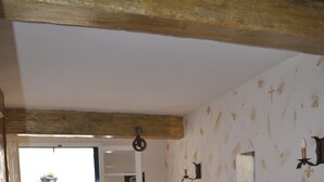 Una sala de estar acogedora con piso de madera, un sofá azul con una manta roja, una pequeña mesa redonda con libros, y una puerta que da a un espacio exterior con una mesa y sillas.