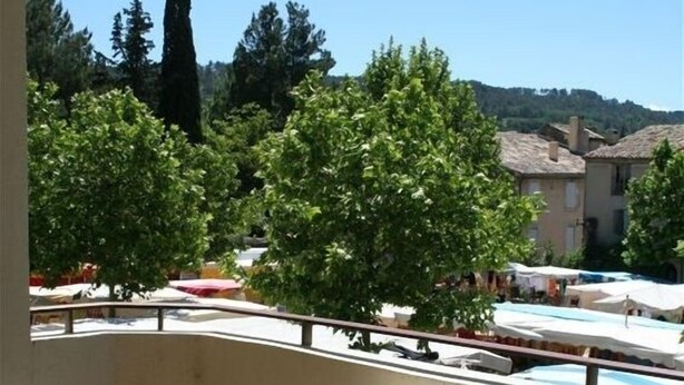 Un balcón con un sofá color crema y un arreglo floral, con vista a una zona de piscina con sombrillas y árboles.