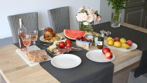 Una mesa de comedor con diversos alimentos, incluyendo un pollo asado, sandía, tomates, manzanas y una barra de pan. También hay dos copas de vino y una botella.