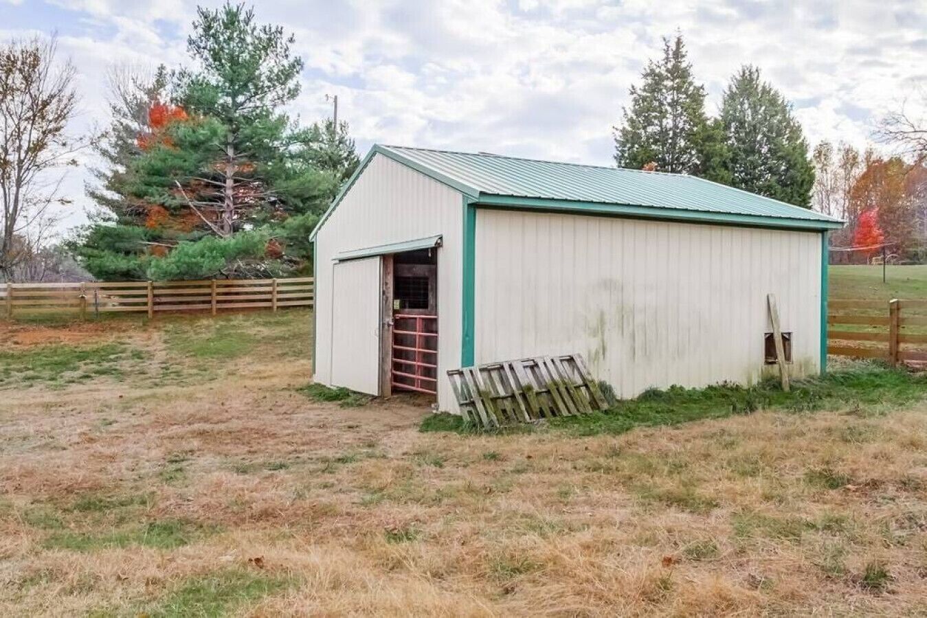 A small, single-story shed with a green roof and white walls, located in a grassy field with trees in the background.