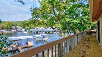 Une terrasse en bois surplombant une marina où sont amarrés des bateaux, un espace couvert avec des sièges, et une vue sur l’eau et les collines en arrière-plan.