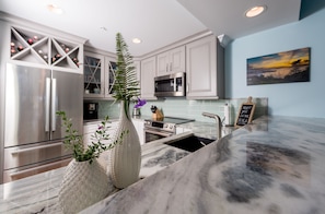 A modern kitchen with white cabinetry, a stainless steel refrigerator, a microwave, and a sink with a marble countertop.