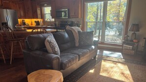 Ein gemütliches Wohnzimmer mit Ledersofa, Couchtisch aus Holz und Küchenzeile, ausgestattet mit Holdekor-Schränken und einem großen Fenster.