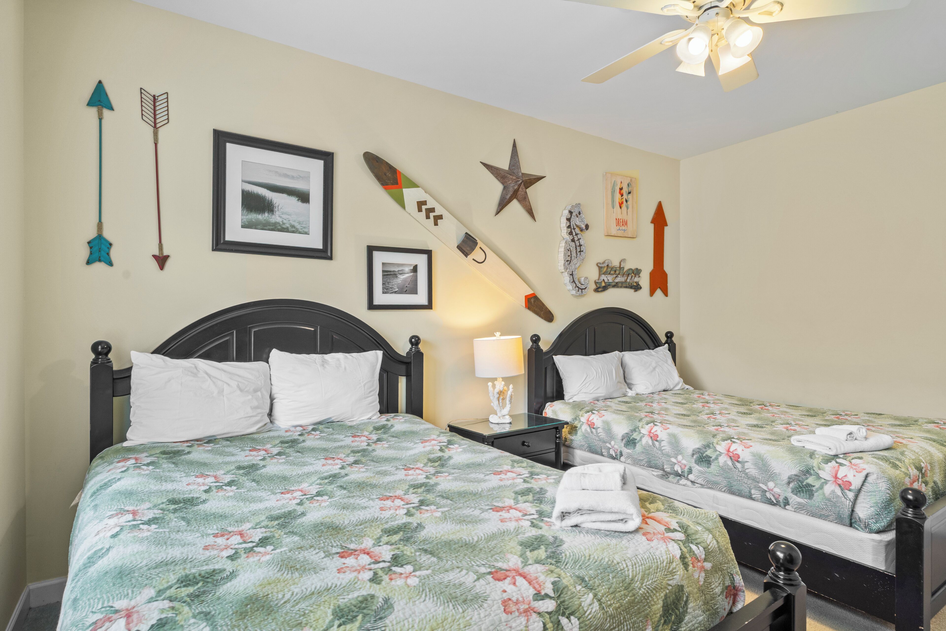 A hotel room with two beds, each with floral bedding, a nightstand with a lamp, and wall decorations including arrows and a star.