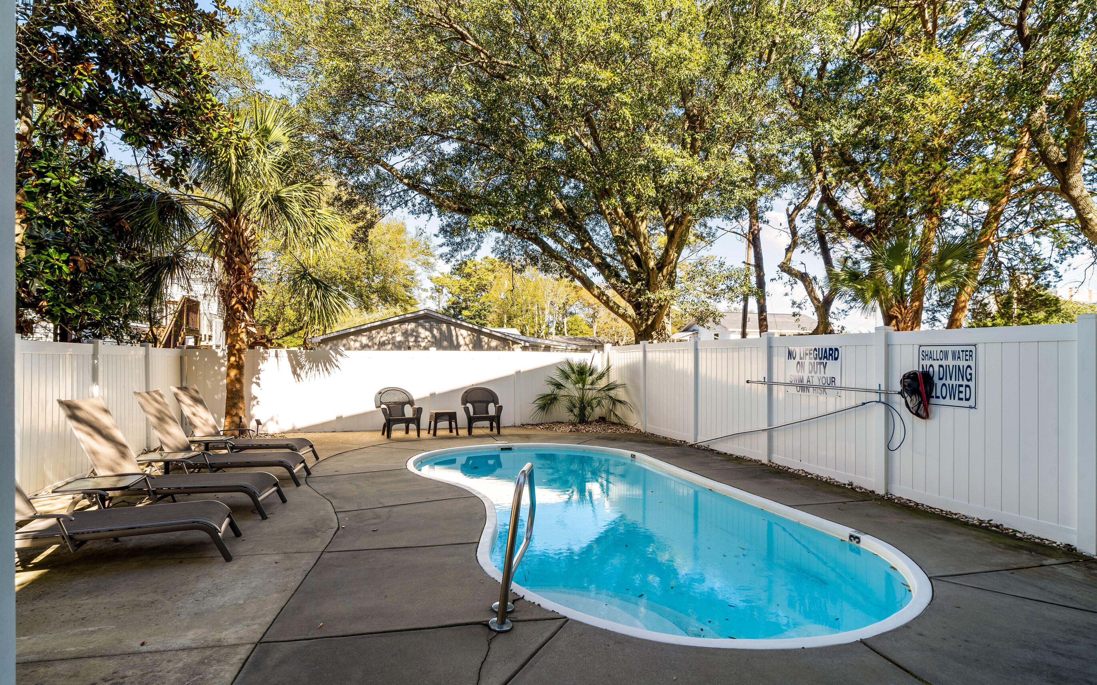 Une piscine entourée d’une clôture blanche, avec des chaises longues et un panneau indiquant l’absence de sauveteur.