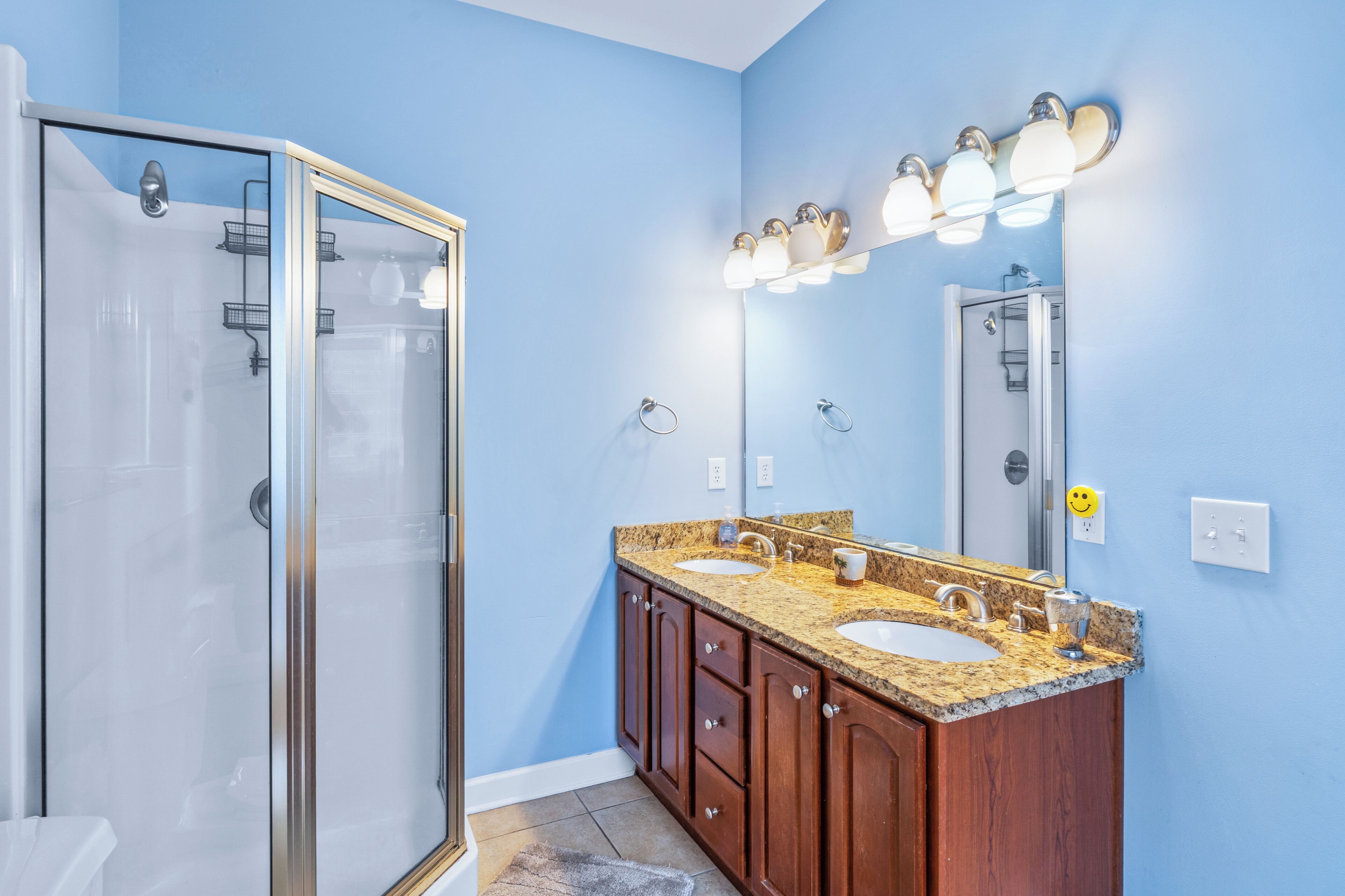 Une salle de bain dotée d’une douche à parois de verre, d’un meuble-lavabo double avec un plan de travail en granit et d’un grand miroir agrémenté de luminaires fixés au mur.