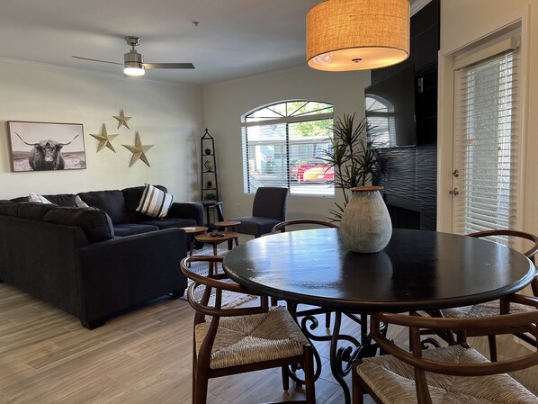 A modern living room with a dark-colored sofa, a round dining table with wicker chairs, a ceiling fan, and a large window with blinds. - Luxury Rental in North Scottsdale-2 Bed/2Bath (Scottsdale)