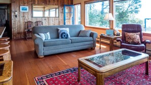 A cozy living room with a wooden ceiling, a grey sofa, a red patterned rug, and a glass coffee table.