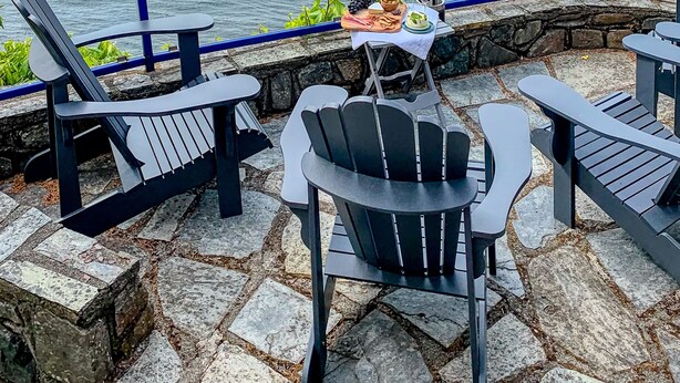 A patio with black Adirondack chairs, a small table with a bottle of wine and a bowl of fruit, and a view of the ocean.