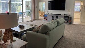 A spacious living room with a large sofa, a flat-screen TV mounted on the wall, and a sliding glass door leading to an outdoor area. - Peaceful Florida Retreat...Spacious 3BR/3BA Private Pool Home (Bonita Springs)