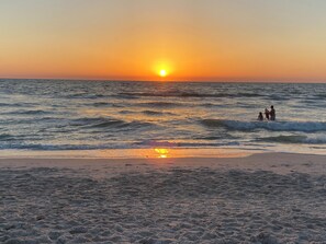 Une plage au coucher du soleil, avec des vagues et quelques personnes dans l’eau.