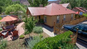 A two-story building with a red roof, surrounded by greenery and a wooden fence. - Moosewood Cottage - Hot Tub, Downtown, Dog Friendly (Moab)