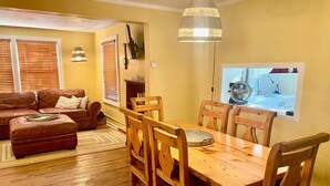 A dining area with a wooden table and chairs, a hanging light fixture, and a window with a view of a kitchen. - Moosewood Cottage - Hot Tub, Downtown, Dog Friendly (Moab)