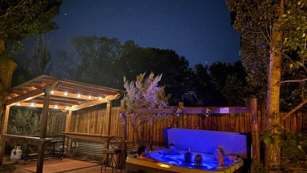 An outdoor hot tub area with a wooden pergola and string lights, surrounded by trees. - Moosewood Cottage - Hot Tub, Downtown, Dog Friendly (Moab)