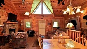A cozy cabin interior with a stone fireplace, wooden walls, a dining table, and a television.