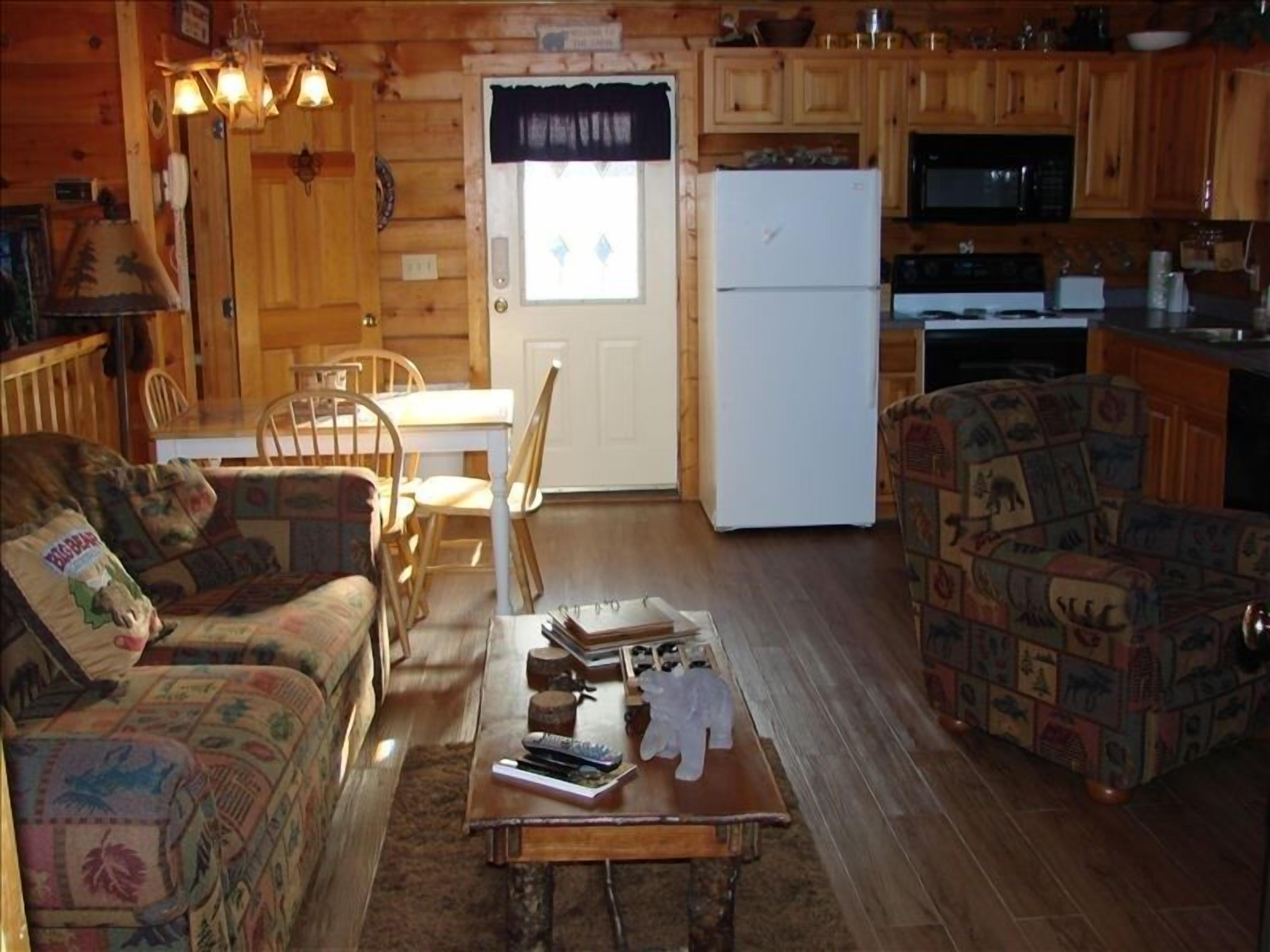 Una habitación acogedora estilo cabaña, con interior de madera, una mesa de comedor con sillas, un sofá, una mesita de centro con libros y un control remoto, y una zona de cocina con refrigerador y microondas.