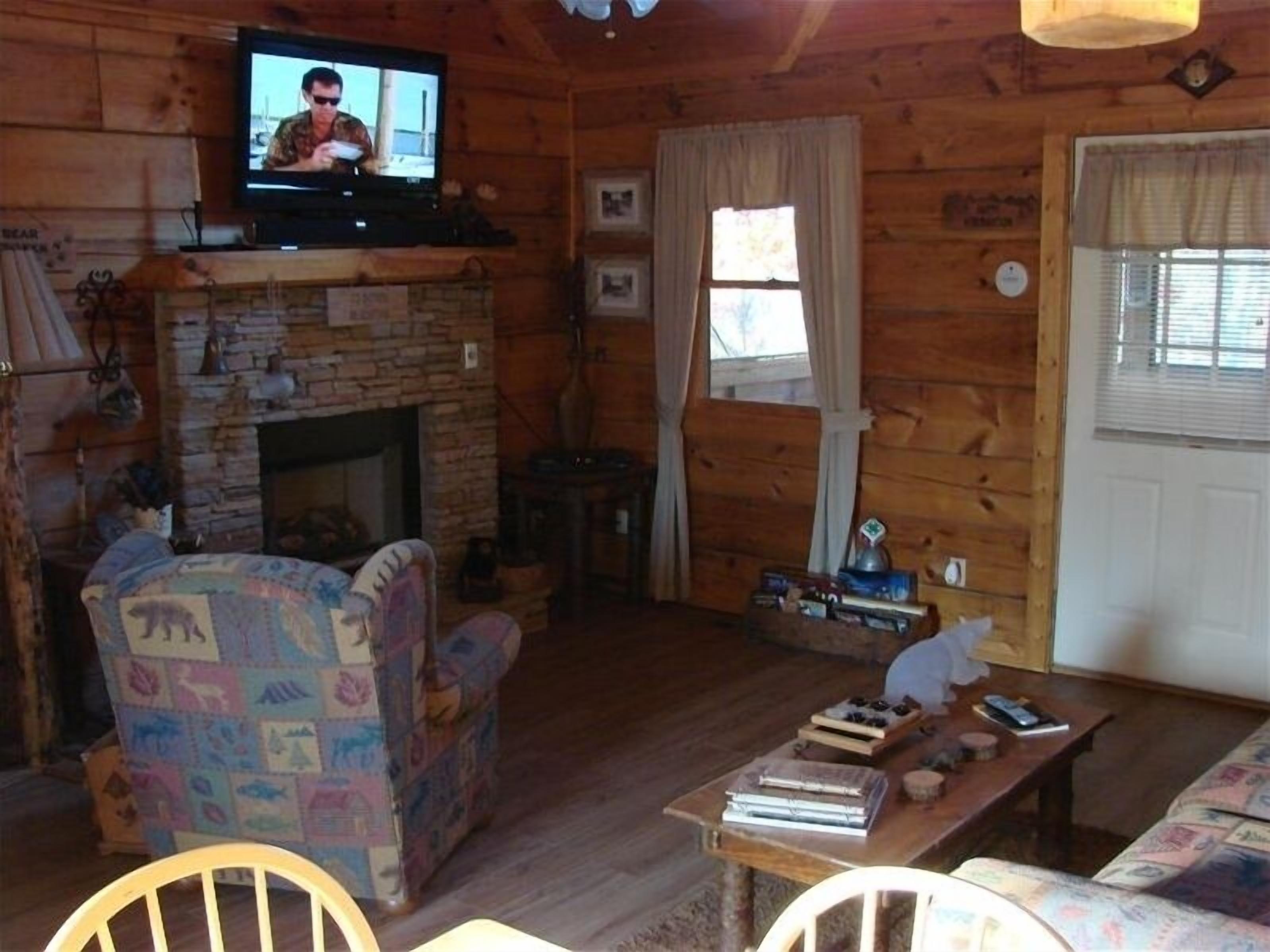 L’intérieur d’un chalet accueillant, avec une cheminée en pierre, un téléviseur fixé au-dessus, un fauteuil à motifs et une table en bois sur laquelle sont posés des livres et une petite figurine.