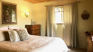 A bedroom with a wooden ceiling, a bed with patterned pillows, a dresser, a wall-mounted lamp, and a window with curtains. - Pet &. Family Treehouse style cabin in woods- Full Time Rental as of Oct 1 (Julian)