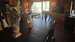 A wooden dining table with a vase and a pyramid-shaped object. There is a stone fireplace, a ceiling fan, and a view of the outdoors through a window. - Pet &. Family Treehouse style cabin in woods- Full Time Rental as of Oct 1 (Julian)