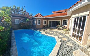 A swimming pool with a stone border, a patio area with seating, and a building with a red-tiled roof.