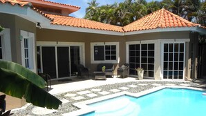 A swimming pool with a tiled deck, surrounded by a house with a tiled roof and large windows.