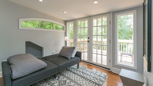 A modern living room with a grey sofa, a patterned rug, and large windows offering a view of a wooden deck.