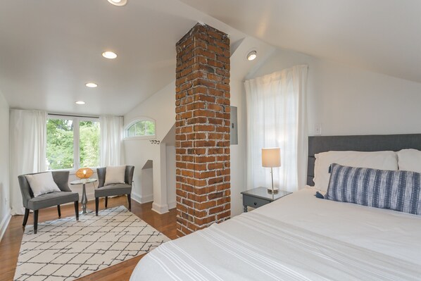 Habitación de hotel con chimenea de ladrillo, una cama grande con ropa blanca, una mesita con lámpara y un sofá con cojines.