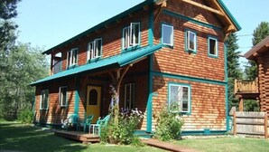 Une cabane en rondins à deux étages, dotée d’une véranda, de nombreuses fenêtres et d’un petit jardin agrémenté de chaises.