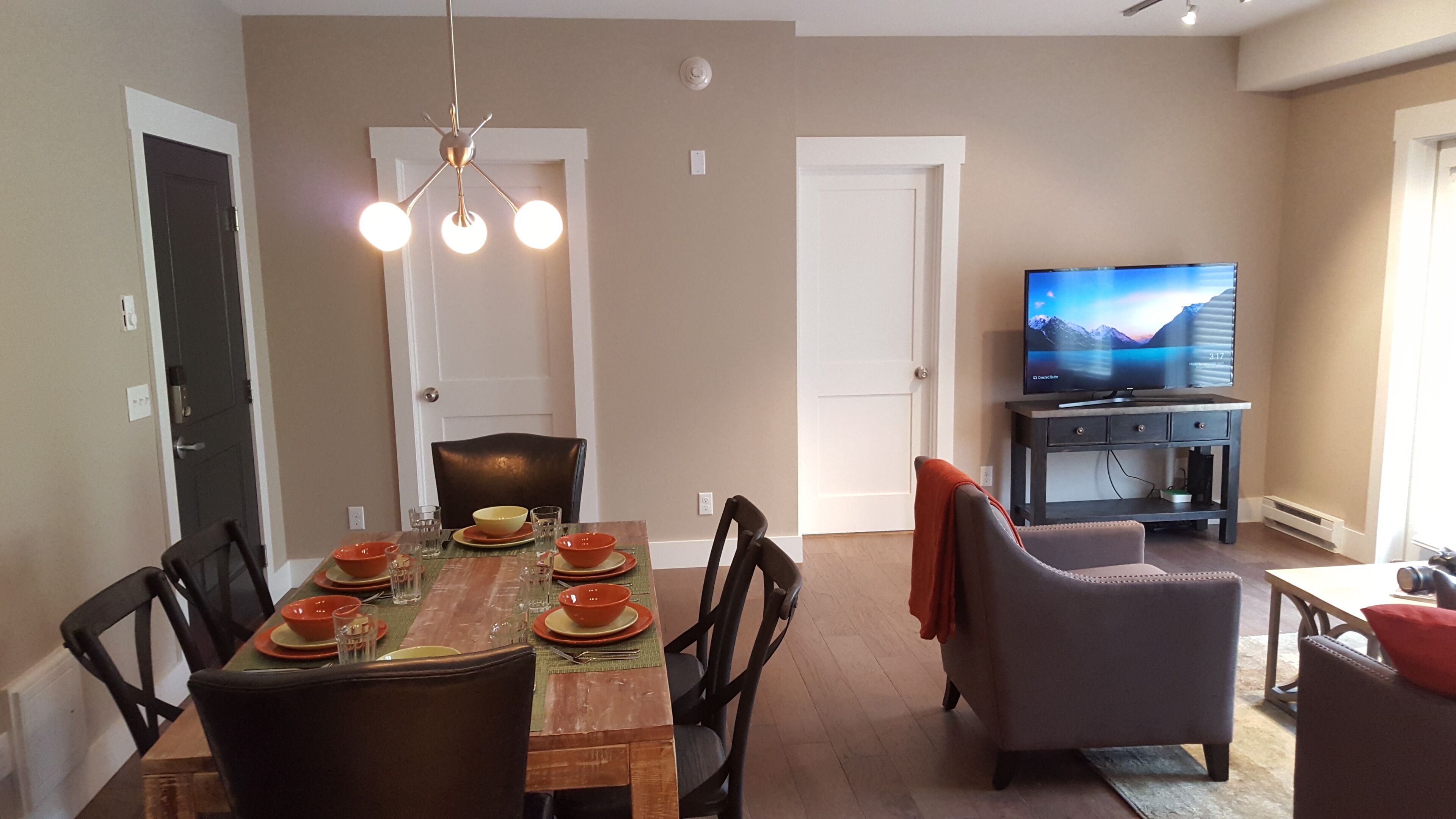 A dining area with a wooden table set for four, surrounded by chairs. There is a television on a stand to the right.