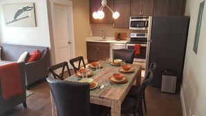 A dining area with a wooden table set for four, surrounded by chairs. A kitchen with modern appliances and a dining table with a placemat is visible in the background. - Top Floor Timbers Condo in Mt. Crested Butte (Crested Butte)