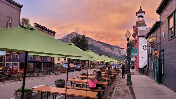 Outdoor wooden tables and benches under green umbrellas, with a mountain backdrop and buildings on either side. - Top Floor Timbers Condo in Mt. Crested Butte (Crested Butte)
