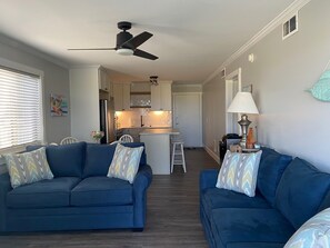 Ein kompakter Wohnbereich mit zwei blauen Sofas, einem Deckenventilator, einer Küchenzeile und einem kleinen Esstisch.