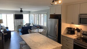 A modern kitchen with a central island, stainless steel appliances, and a dining area with a blue sofa and white chairs. - Oceanfront 2BR/2Bath Condo in Carolina Beach NC  (Carolina Beach)