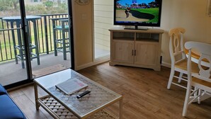 A room with a television displaying a golf event, a wooden cabinet, a glass coffee table, a white chair, and a view of the outdoors through a sliding door. - Ocean Front 1st Row ~ Private Corner Unit ~ Close to Pool, Beach Bar & Boardwalk (Hilton Head Island)