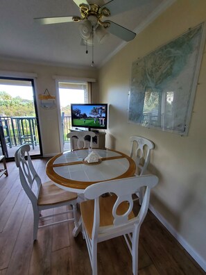 A dining area with a round table, white chairs, a ceiling fan, and a television displaying a golf game. - Ocean Front 1st Row ~ Private Corner Unit ~ Close to Pool, Beach Bar & Boardwalk (Hilton Head Island)