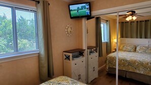 A bedroom with a bed, a TV mounted on the wall, a bedside table, and a window with blinds. - Ocean Front 1st Row ~ Private Corner Unit ~ Close to Pool, Beach Bar & Boardwalk (Hilton Head Island)