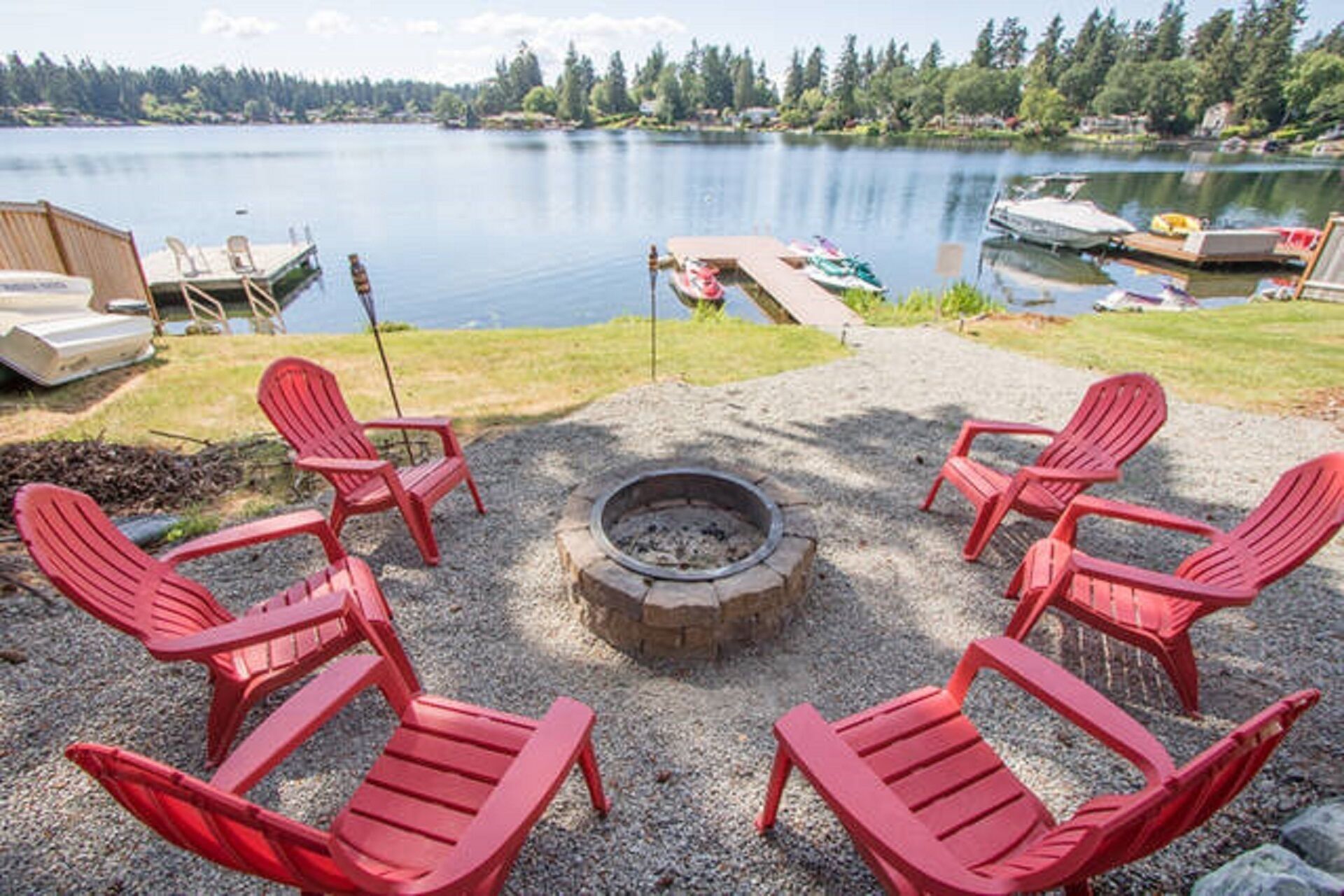 A lakeside deck with red Adirondack chairs, a fire pit, and boats docked in the background.