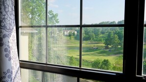 A view through a window with a floral curtain, revealing a green landscape with trees and a building in the distance. - Top-Floor Branson Condo w/ Stunning Views & 2 King Beds (Branson)