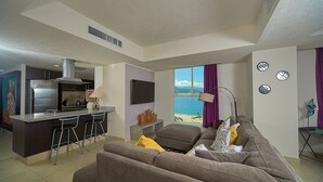 Una sala de estar moderna con un sofá grande, una zona de cocina con refrigerador y taburetes de bar, y vista al océano a través de un ventanal amplio.