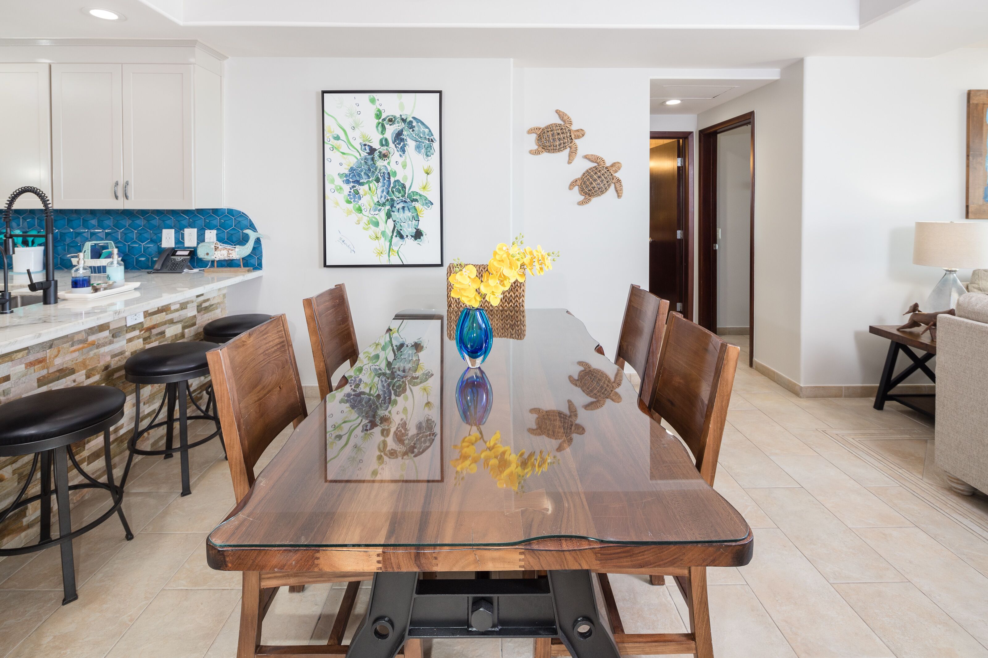 A dining area with a glass-top table, wooden chairs, and a vase with yellow flowers. There is a kitchen in the background with white cabinets and a blue backsplash.