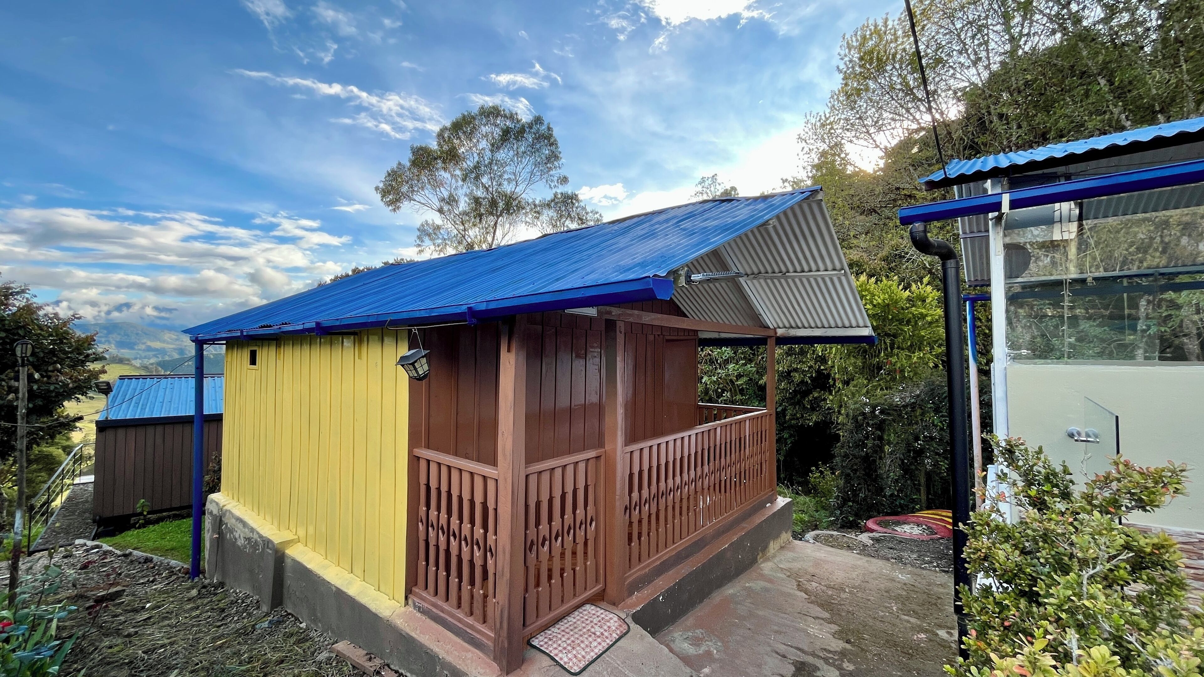 A small wooden structure with a blue roof and a yellow side, featuring a wooden railing and a small porch area.
